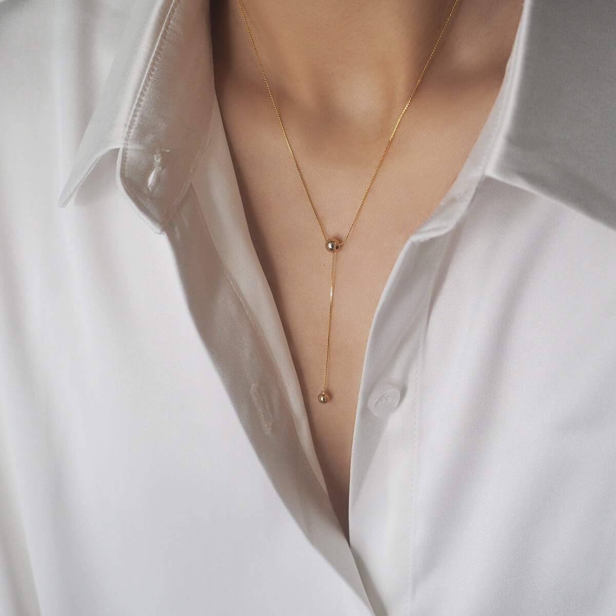 A woman in a white shirt models a beautiful gold necklace featuring a double chain and ball pendant, enhancing her outfit's elegance.