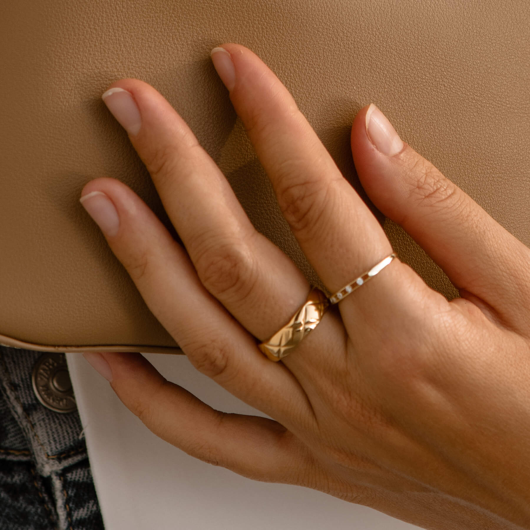 A close-up of a woman's hand gripping a tan purse, featuring a striking gold ring adorned with a unique "X" design.