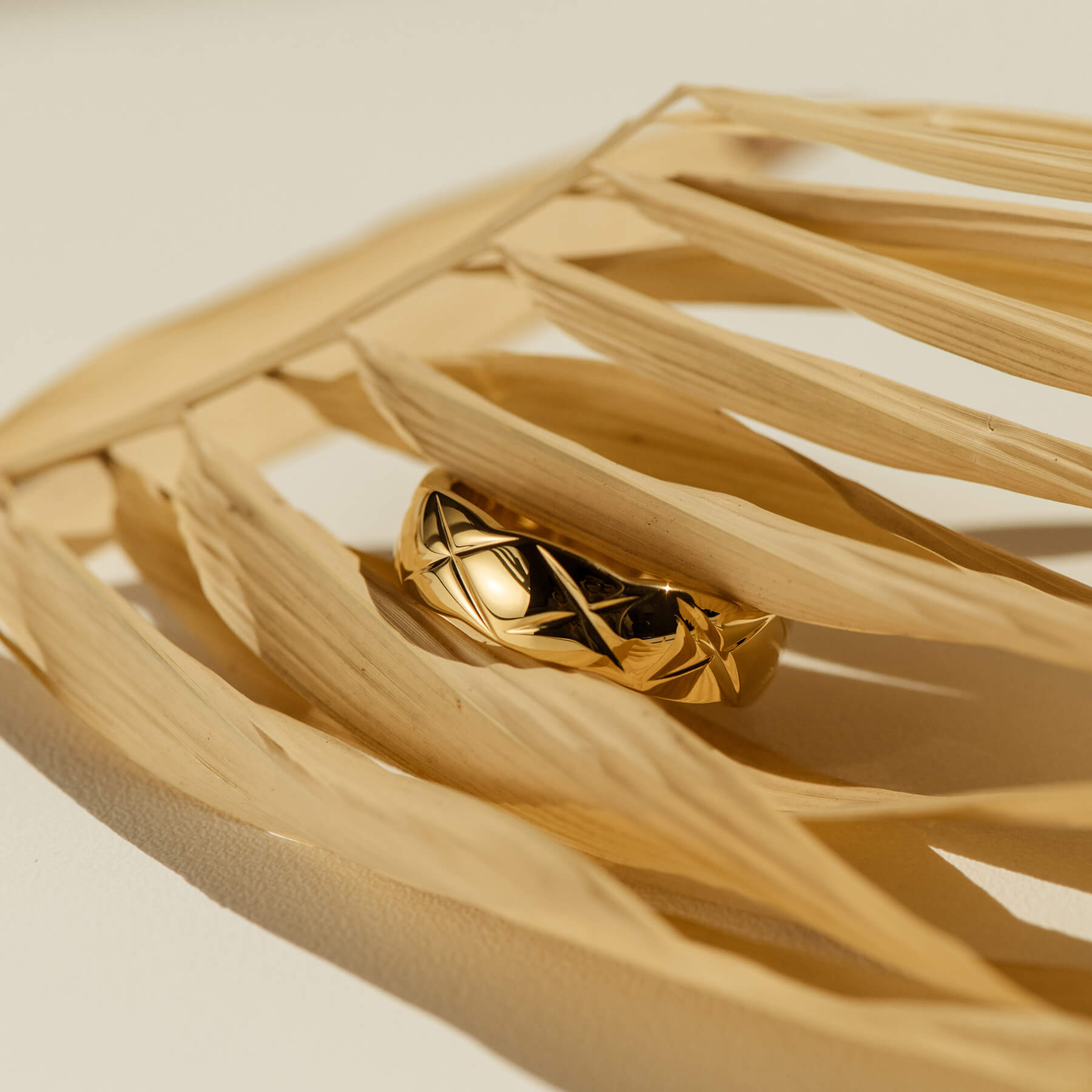 A stunning gold ring featuring an intricate "X" design lies atop a green palm leaf, highlighting its exquisite craftsmanship.